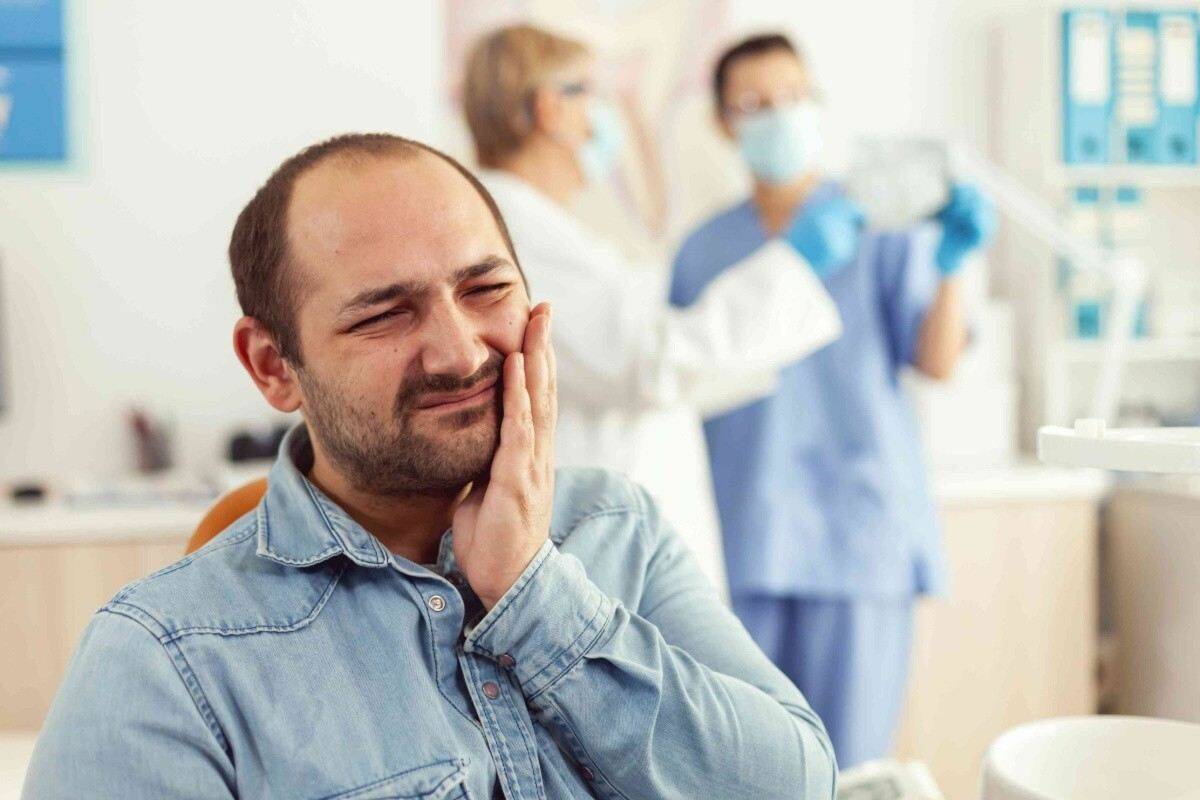 Man holding his cheek in pain while seated in an orthodontic clinic with dental staff in background.